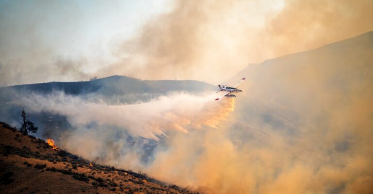 A firefighting plane combats a wildfire, spreading water over rugged terrain at sunset.