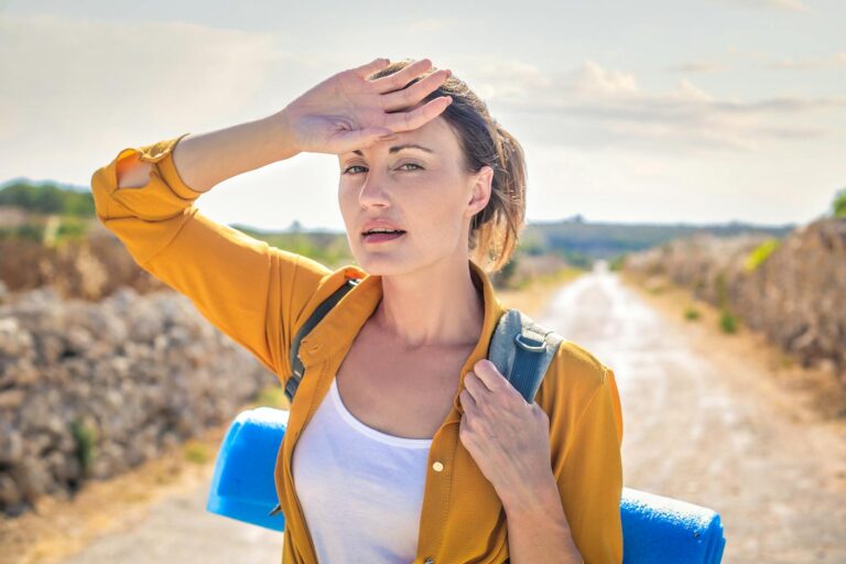 A woman stands on a sunny pathway outdoors with a backpack, enjoying a summer day.